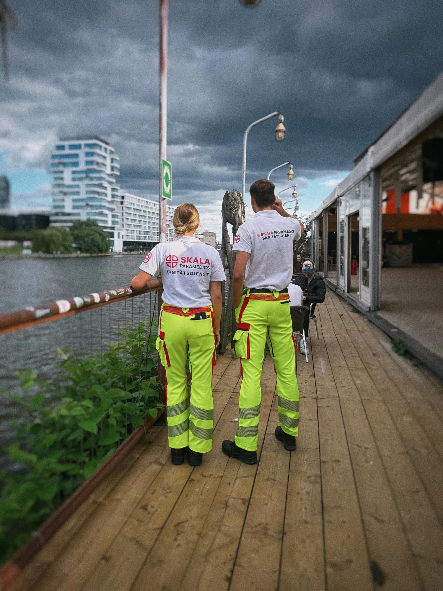 Zwei Sanitäter:innen von SKALA Paramedics von hinten auf einem Holzsteg an der Spree